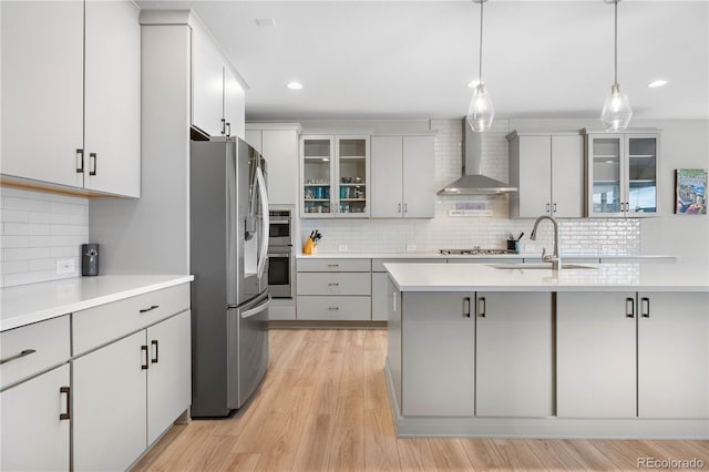 kitchen featuring decorative light fixtures, light countertops, wall chimney range hood, appliances with stainless steel finishes, and glass insert cabinets