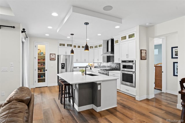 kitchen with decorative light fixtures, an island with sink, sink, stainless steel appliances, and wall chimney range hood