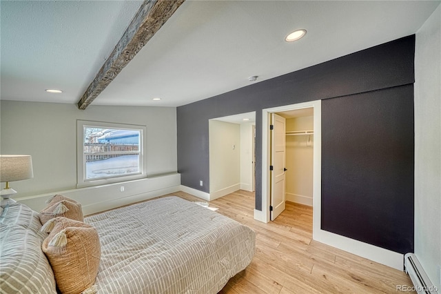 bedroom featuring a walk in closet, baseboards, beamed ceiling, baseboard heating, and light wood-style flooring