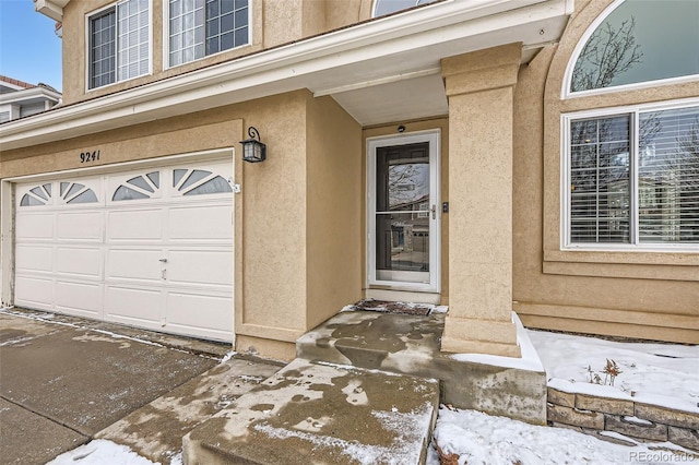 snow covered property entrance with an attached garage and stucco siding