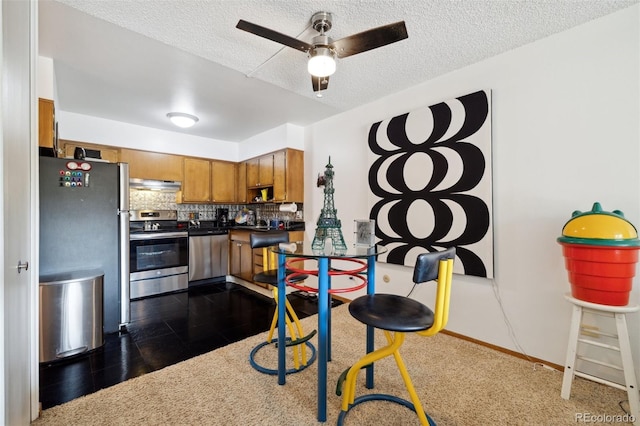 kitchen featuring a textured ceiling, under cabinet range hood, appliances with stainless steel finishes, brown cabinets, and tasteful backsplash