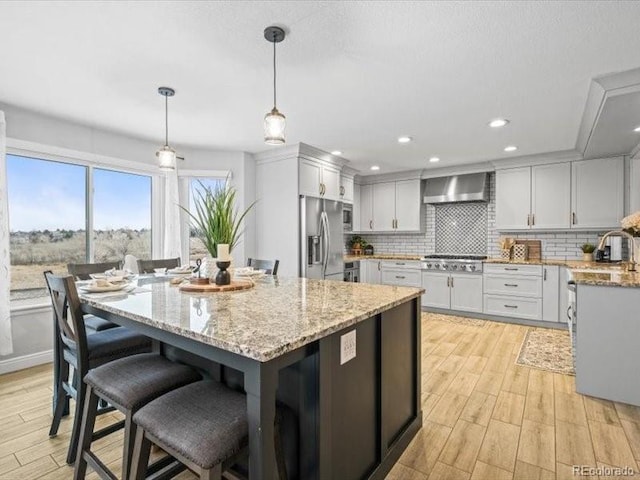 kitchen with a center island with sink, white cabinets, wall chimney exhaust hood, stainless steel appliances, and pendant lighting