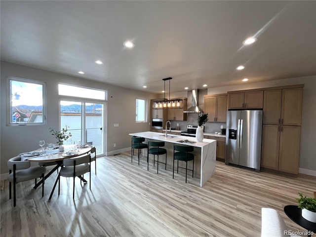 kitchen with a center island with sink, wall chimney range hood, sink, hanging light fixtures, and stainless steel appliances