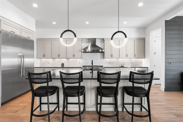 kitchen featuring decorative backsplash, an island with sink, built in refrigerator, light wood-type flooring, and wall chimney range hood