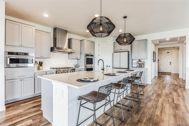 kitchen featuring gray cabinets, wall chimney range hood, a kitchen island with sink, and tasteful backsplash