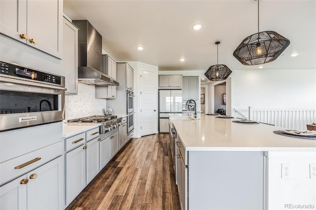 kitchen with wall chimney range hood, an island with sink, pendant lighting, gray cabinets, and appliances with stainless steel finishes