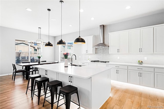 kitchen with backsplash, a kitchen island with sink, white cabinets, wall chimney range hood, and hanging light fixtures