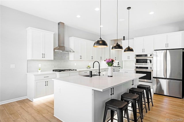 kitchen featuring a center island with sink, sink, wall chimney exhaust hood, appliances with stainless steel finishes, and decorative light fixtures