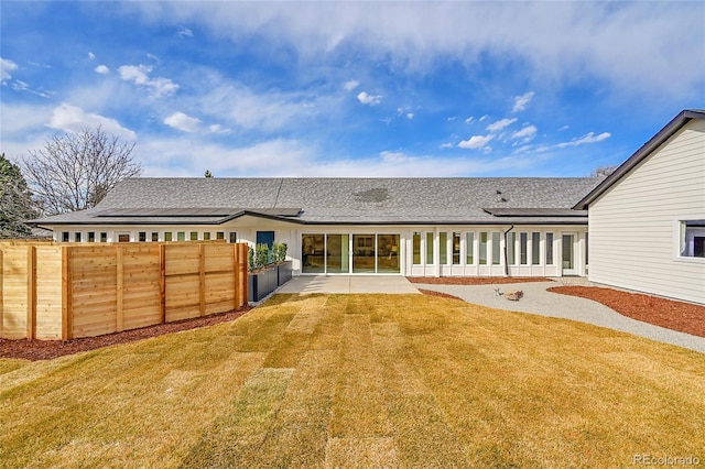 back of property with fence, roof with shingles, a patio area, a lawn, and roof mounted solar panels