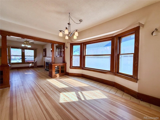 unfurnished dining area featuring light wood-type flooring, ceiling fan with notable chandelier, a textured ceiling, baseboards, and a brick fireplace