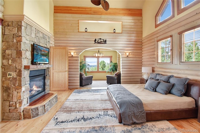 bedroom featuring a notable chandelier, a stone fireplace, light hardwood / wood-style floors, a towering ceiling, and rustic walls