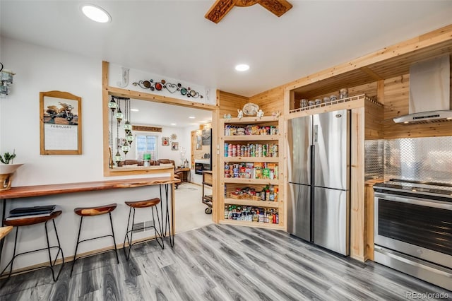 kitchen featuring wall chimney range hood, hardwood / wood-style flooring, backsplash, stainless steel appliances, and a kitchen breakfast bar