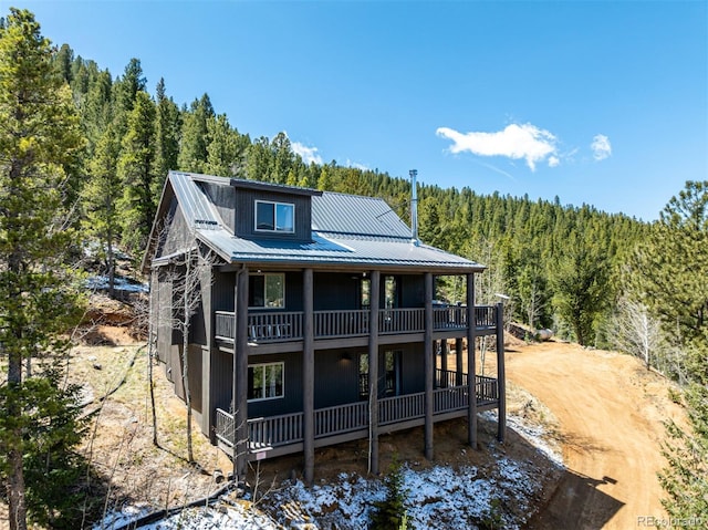 back of house with a view of trees, a balcony, and metal roof