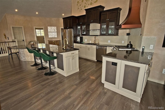 kitchen with stainless steel appliances, a breakfast bar area, custom exhaust hood, a kitchen island with sink, and dark hardwood / wood-style flooring