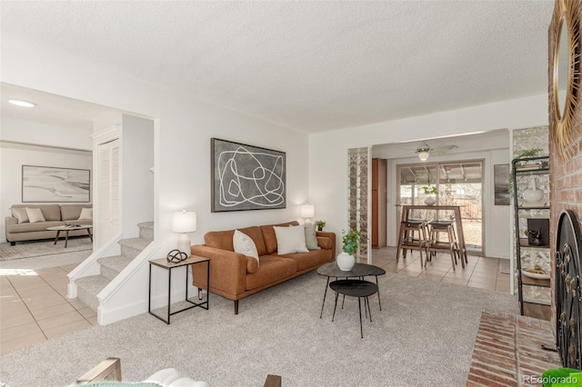 living room featuring ceiling fan, stairway, a textured ceiling, a brick fireplace, and light tile patterned flooring