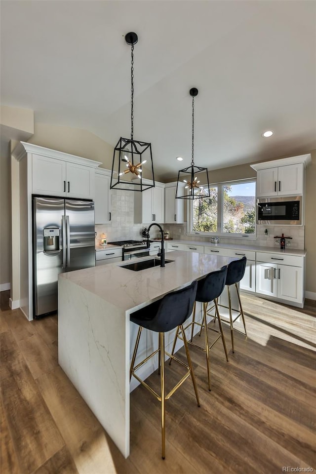 kitchen with a sink, an island with sink, wood finished floors, built in microwave, and stainless steel fridge