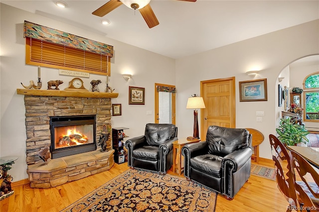 living room with a stone fireplace, ceiling fan, light wood finished floors, and arched walkways