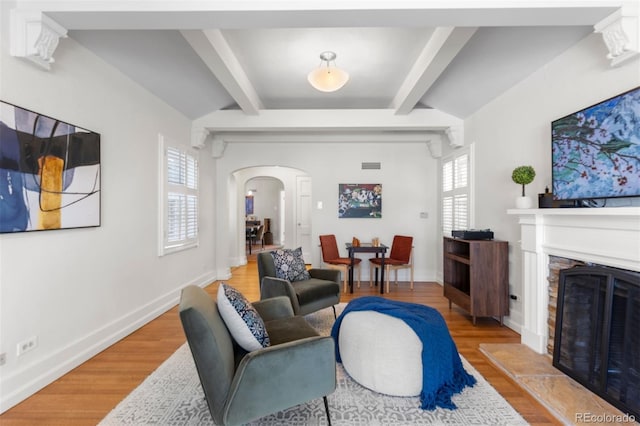 living area featuring arched walkways, wood finished floors, a fireplace with raised hearth, baseboards, and beamed ceiling