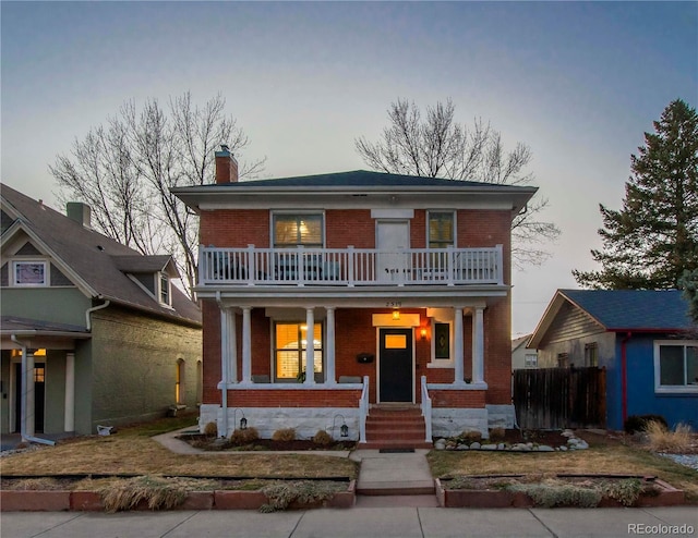 view of front facade featuring a balcony, covered porch, brick siding, fence, and a chimney