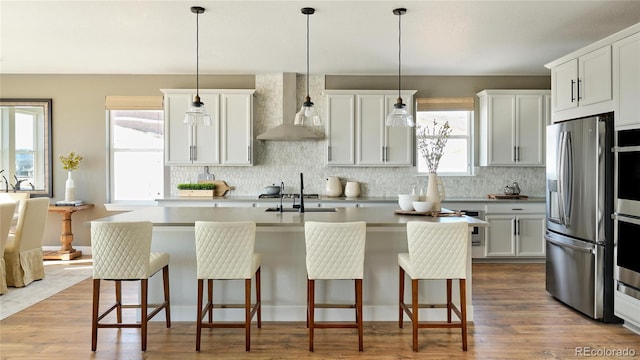 kitchen with hanging light fixtures, wall chimney exhaust hood, an island with sink, appliances with stainless steel finishes, and white cabinetry