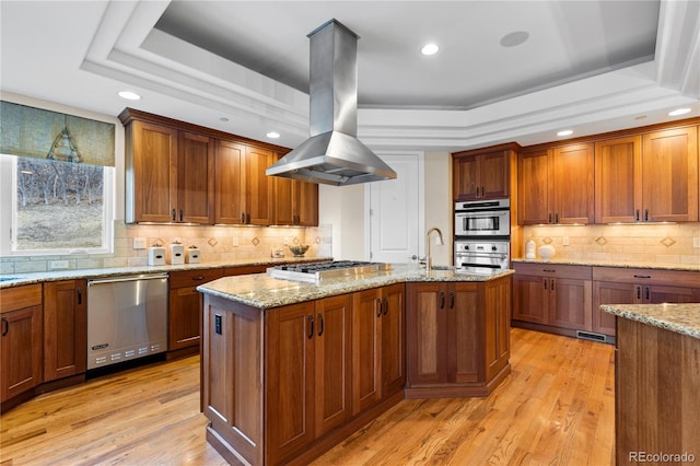 kitchen with stainless steel appliances, an island with sink, light stone countertops, island range hood, and a tray ceiling