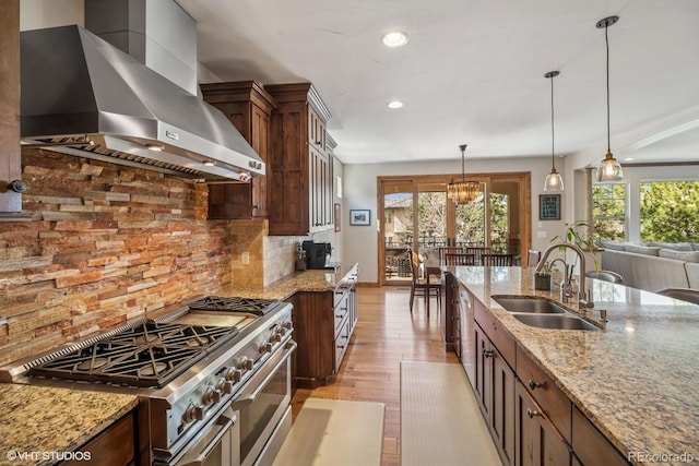 kitchen with a sink, hanging light fixtures, light wood-type flooring, wall chimney exhaust hood, and appliances with stainless steel finishes