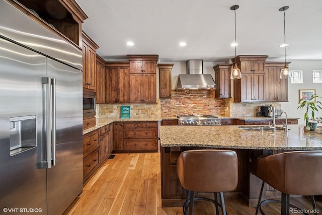 kitchen with brown cabinets, a sink, wall chimney exhaust hood, light wood-style flooring, and built in appliances