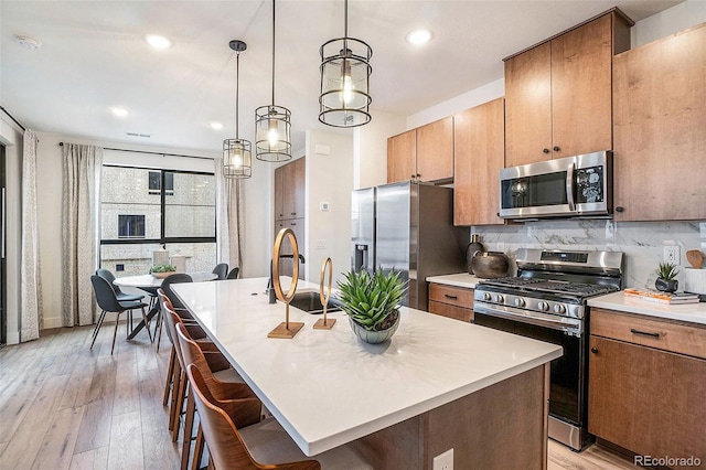 kitchen with an island with sink, light wood-type flooring, decorative light fixtures, and appliances with stainless steel finishes