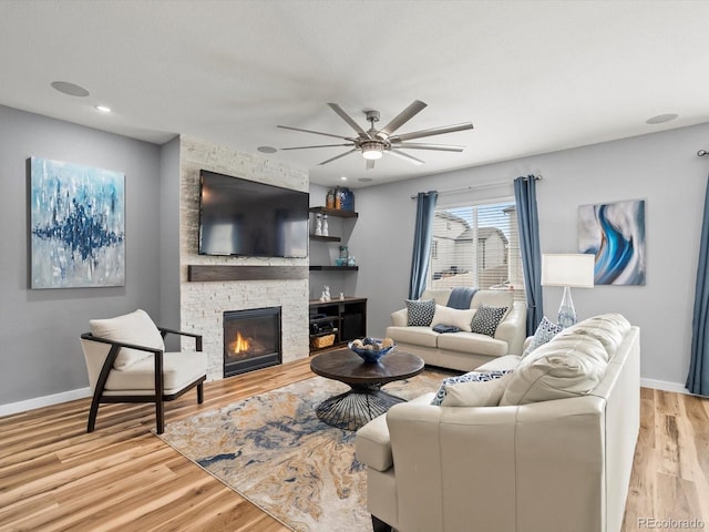 living area with a stone fireplace, light wood-style flooring, a ceiling fan, and baseboards
