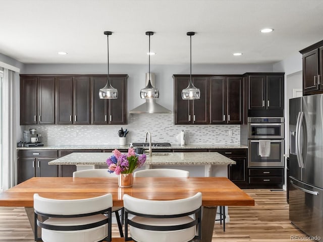 kitchen with light wood-type flooring, wall chimney range hood, backsplash, dark brown cabinets, and stainless steel appliances