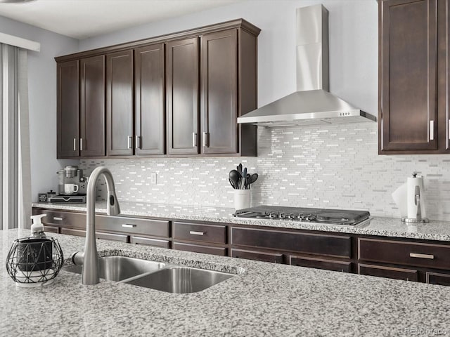 kitchen featuring tasteful backsplash, dark brown cabinetry, light stone counters, wall chimney range hood, and a sink