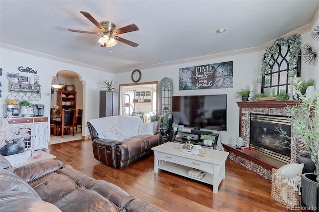 living room with arched walkways, a textured ceiling, a stone fireplace, wood finished floors, and ornamental molding