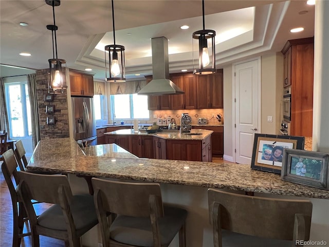 kitchen featuring kitchen peninsula, a tray ceiling, island range hood, pendant lighting, and stainless steel appliances