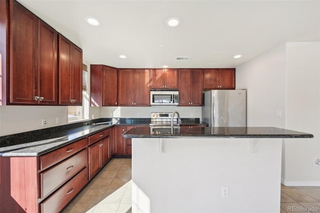 kitchen with dark stone countertops, an island with sink, light tile patterned floors, and appliances with stainless steel finishes