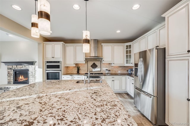 kitchen featuring decorative backsplash, light stone counters, stainless steel appliances, wall chimney range hood, and hanging light fixtures