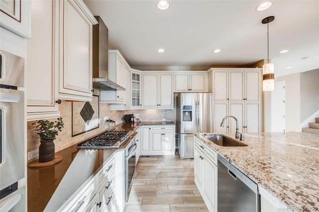 kitchen featuring sink, hanging light fixtures, wall chimney range hood, light stone counters, and appliances with stainless steel finishes