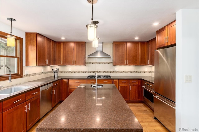 kitchen featuring stainless steel appliances, wall chimney range hood, a center island with sink, a sink, and decorative light fixtures