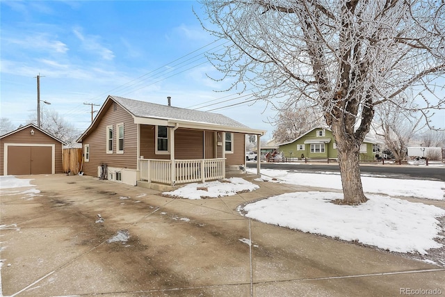 snow covered property featuring concrete driveway, a detached garage, an outbuilding, a shed, and a porch