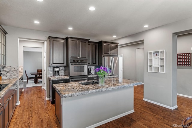 1609 S Catawba Cir, Aurora, CO 80018 - kitchen featuring appliances with stainless steel finishes, backsplash, dark wood-style floors, a center island with sink, and light stone countertops