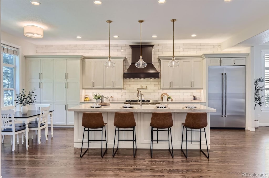 kitchen featuring decorative light fixtures, custom exhaust hood, a kitchen island with sink, stainless steel appliances, and dark hardwood / wood-style flooring