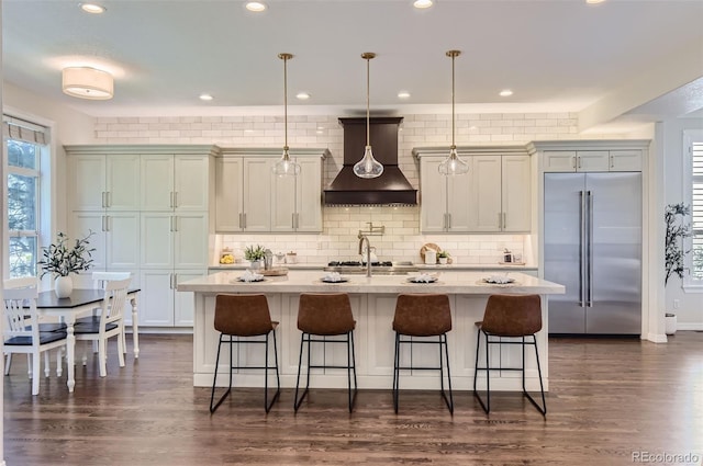 kitchen featuring decorative light fixtures, custom exhaust hood, a kitchen island with sink, stainless steel appliances, and dark hardwood / wood-style flooring