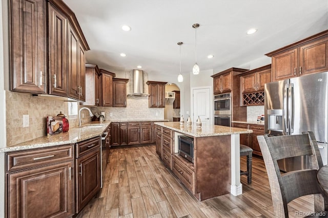 kitchen with stainless steel appliances, hanging light fixtures, a sink, a kitchen island, and wall chimney exhaust hood