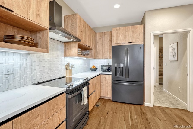 kitchen with light stone countertops, appliances with stainless steel finishes, wall chimney range hood, light brown cabinets, and light hardwood / wood-style flooring