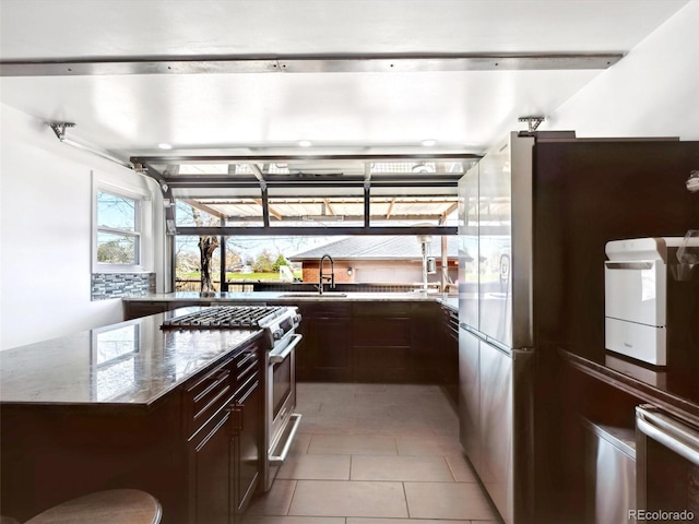 kitchen featuring stainless steel appliances, dark brown cabinets, light tile patterned floors, a sink, and light stone countertops
