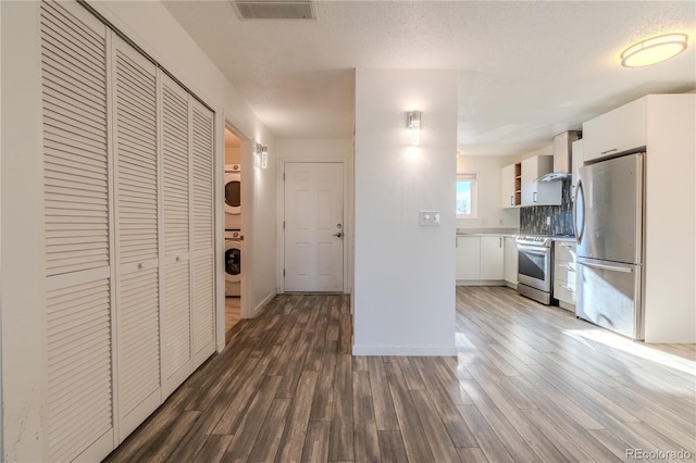 kitchen with appliances with stainless steel finishes, backsplash, stacked washing maching and dryer, wall chimney exhaust hood, and white cabinetry