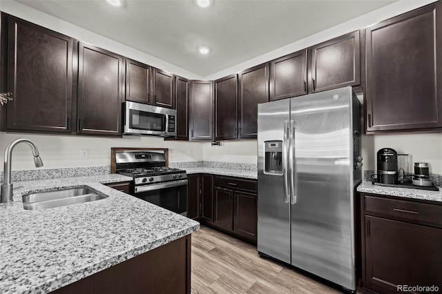 kitchen with sink, dark brown cabinetry, stainless steel appliances, light stone countertops, and light wood-type flooring