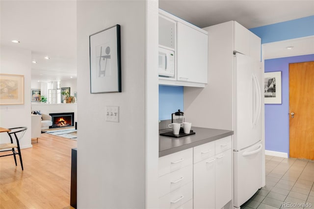 kitchen featuring dark countertops, baseboards, a warm lit fireplace, white cabinets, and white appliances