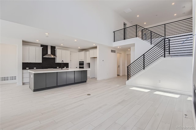 kitchen with tasteful backsplash, a center island with sink, wall chimney exhaust hood, white cabinetry, and light wood-type flooring