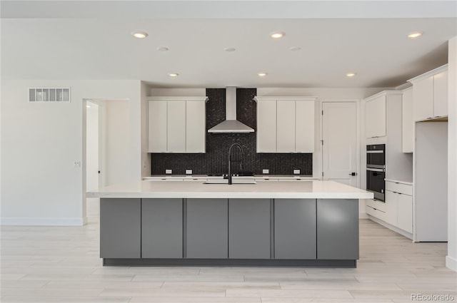 kitchen featuring wall chimney exhaust hood, white cabinets, a center island with sink, and double oven