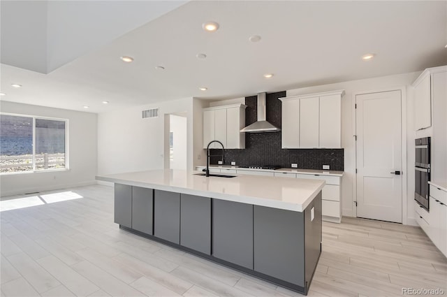 kitchen with white cabinets, sink, tasteful backsplash, a kitchen island with sink, and wall chimney exhaust hood